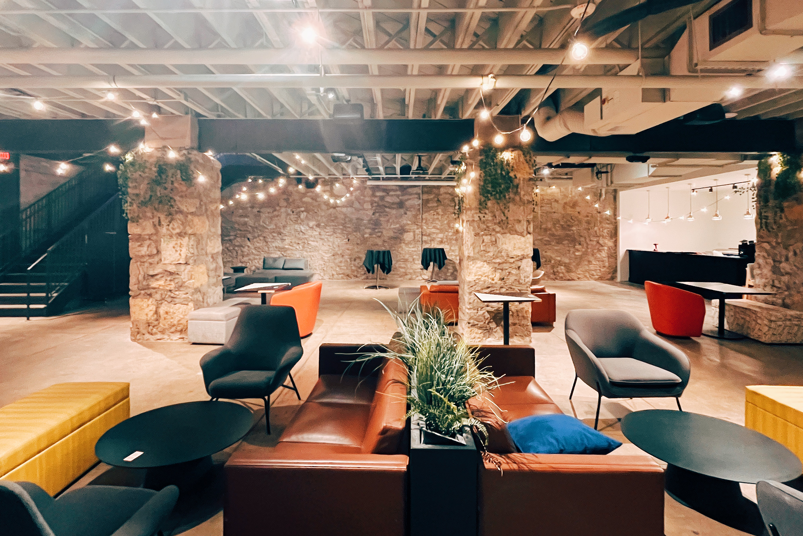 Looking across an entertaining cellar space decorated with brow leather couches and brightly colored seating options. The space is lit from string lighting in the rafters.