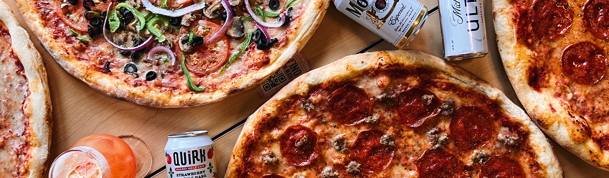 Three pizzas; pepperoni and sausage, pepperoni, and veggie sit on wooden table with beers and a peach cocktail in between.
